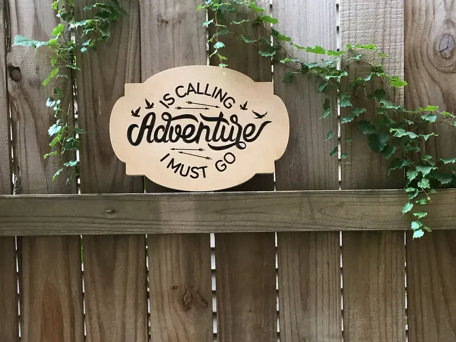 Finished wood sign reading Adventure Must Go displayed on a garden fence