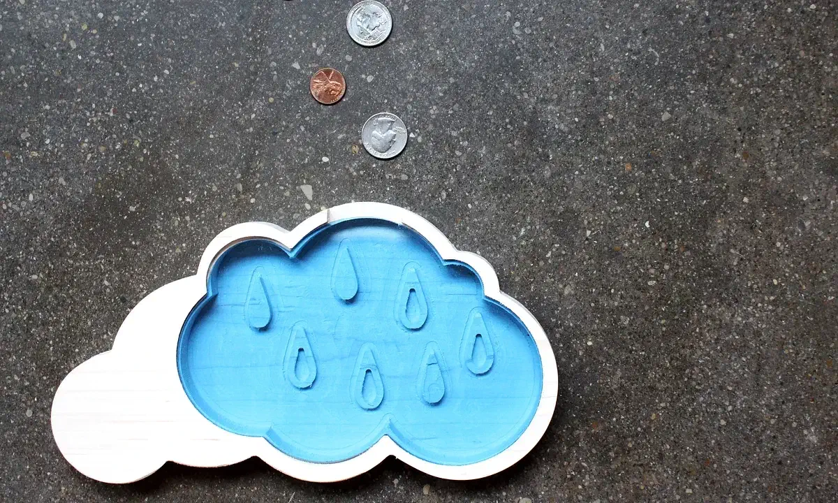 A blue cloud-shaped coin bank with a white border and raindrops, photographed with coins on a dark surface