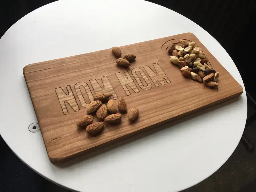 A finished wooden board with NOM NOM lettering and an almond-shaped recessed dish