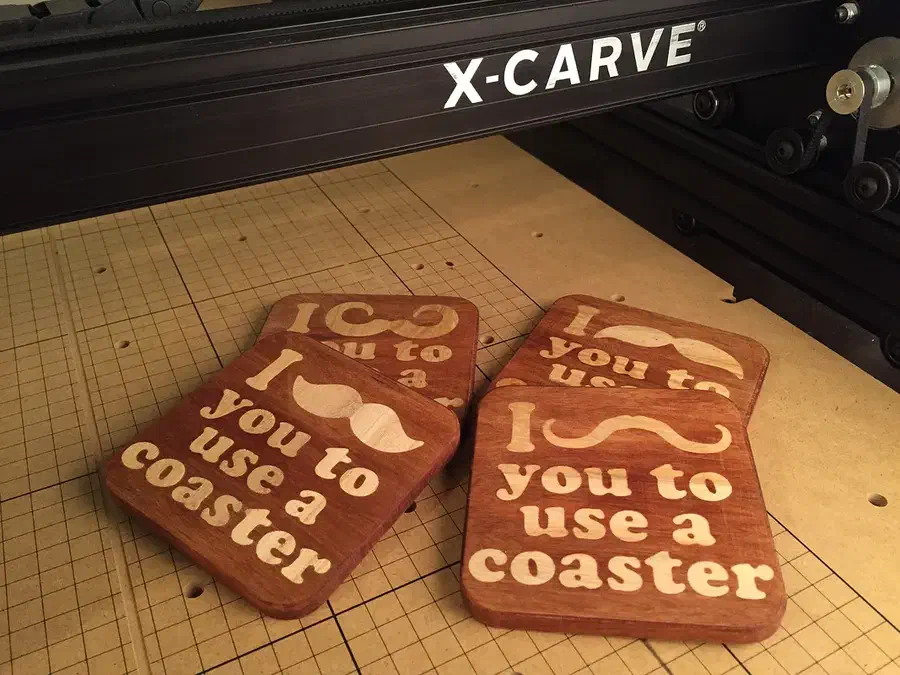 Four rectangular wooden coasters with contrasting pale mustache designs and lettering on an X-Carve bed