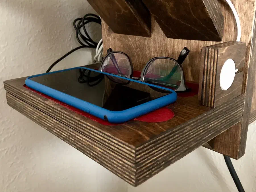 A stained plywood charging tray on a shelf holding a blue-cased phone and eyeglasses beside a wooden watch-charger support