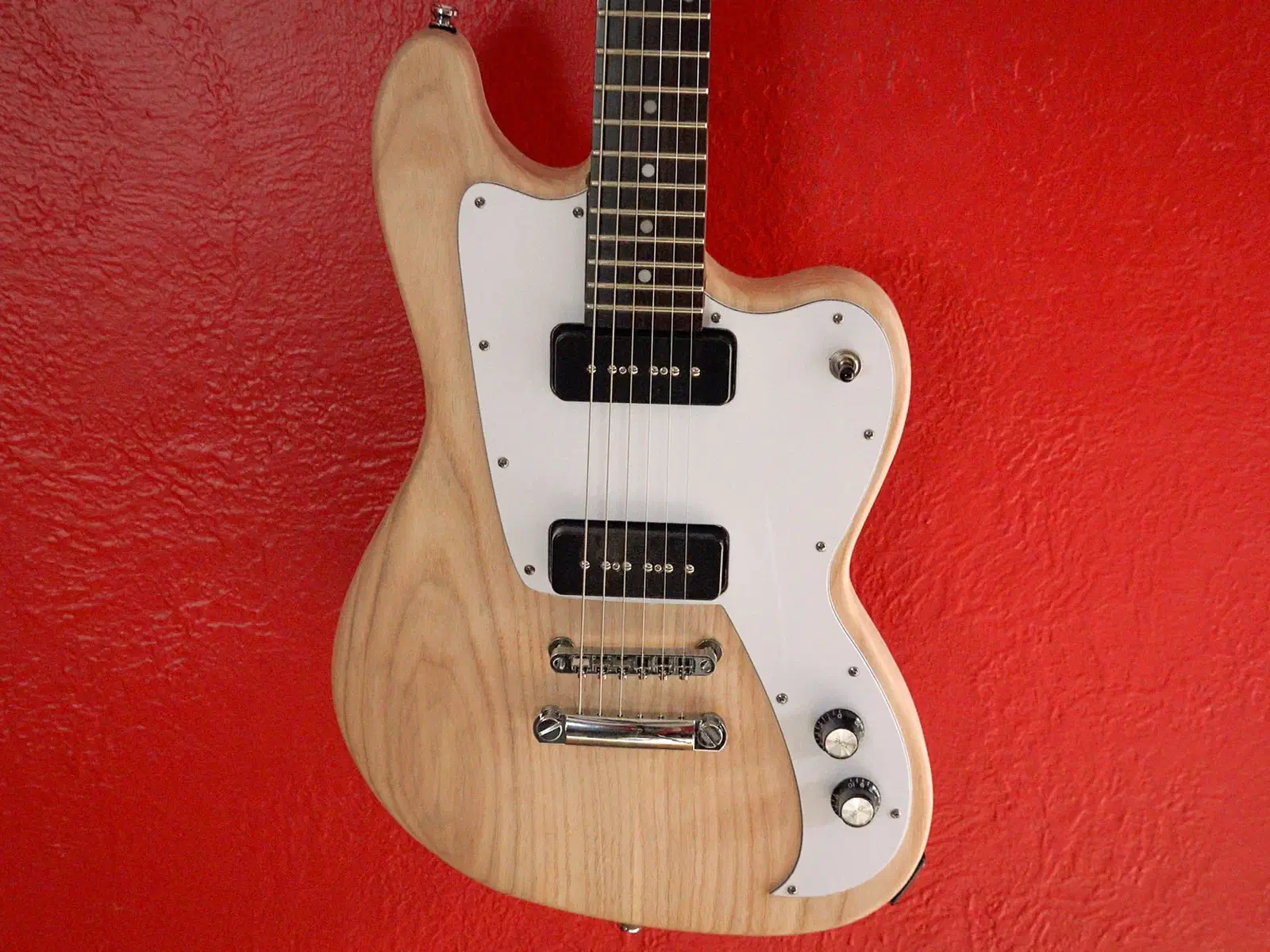 A natural-wood electric guitar body with a white pickguard, black pickups and metal bridge against a red wall