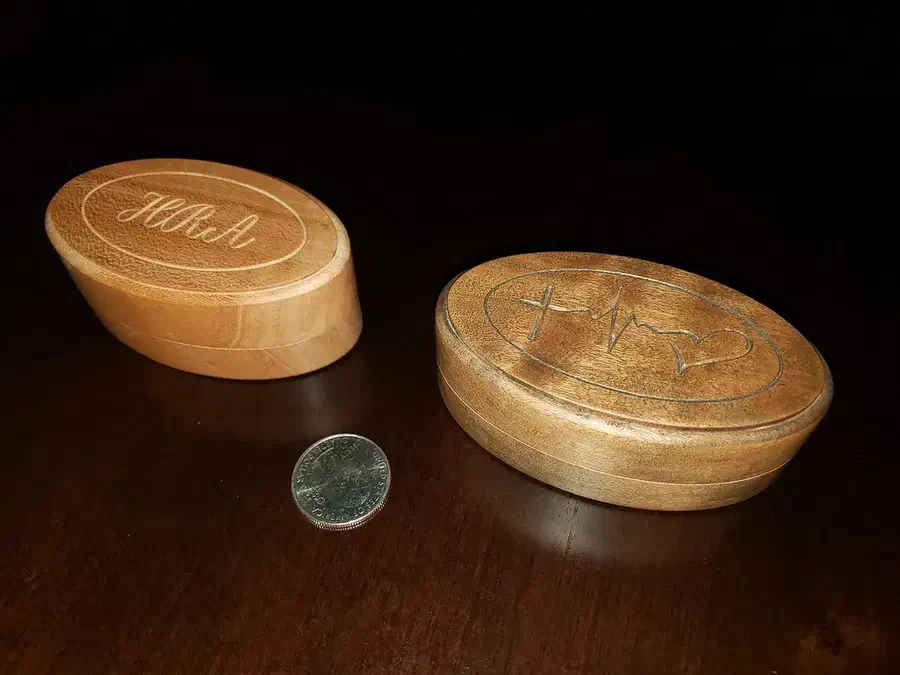 Two small oval wooden ring boxes with engraved lids beside a coin for scale
