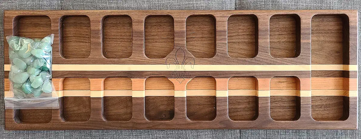 Walnut Mancala board with rows of rectangular pockets, light wood inlay strips, and a bag of game stones resting over one end