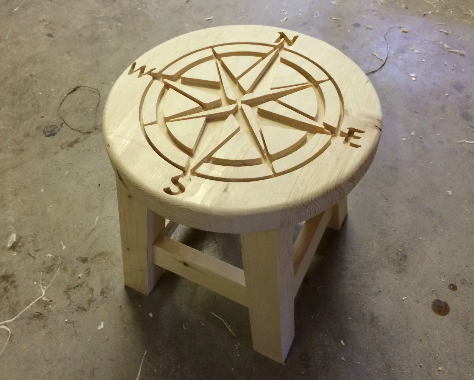 A round wooden step stool with a carved compass rose on its pale top and four legs with stretchers