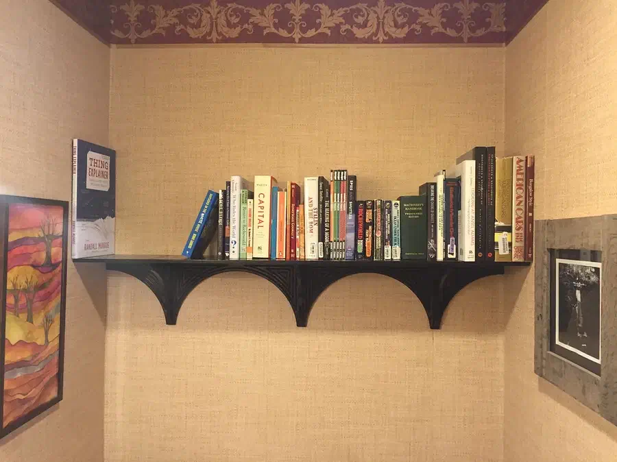 A long dark wooden shelf holding books in a corner above a tiled wall