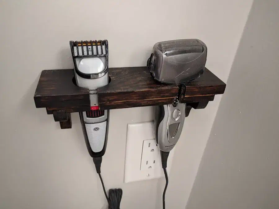 A dark wooden wall shelf holding an electric razor and beard trimmer above a power outlet